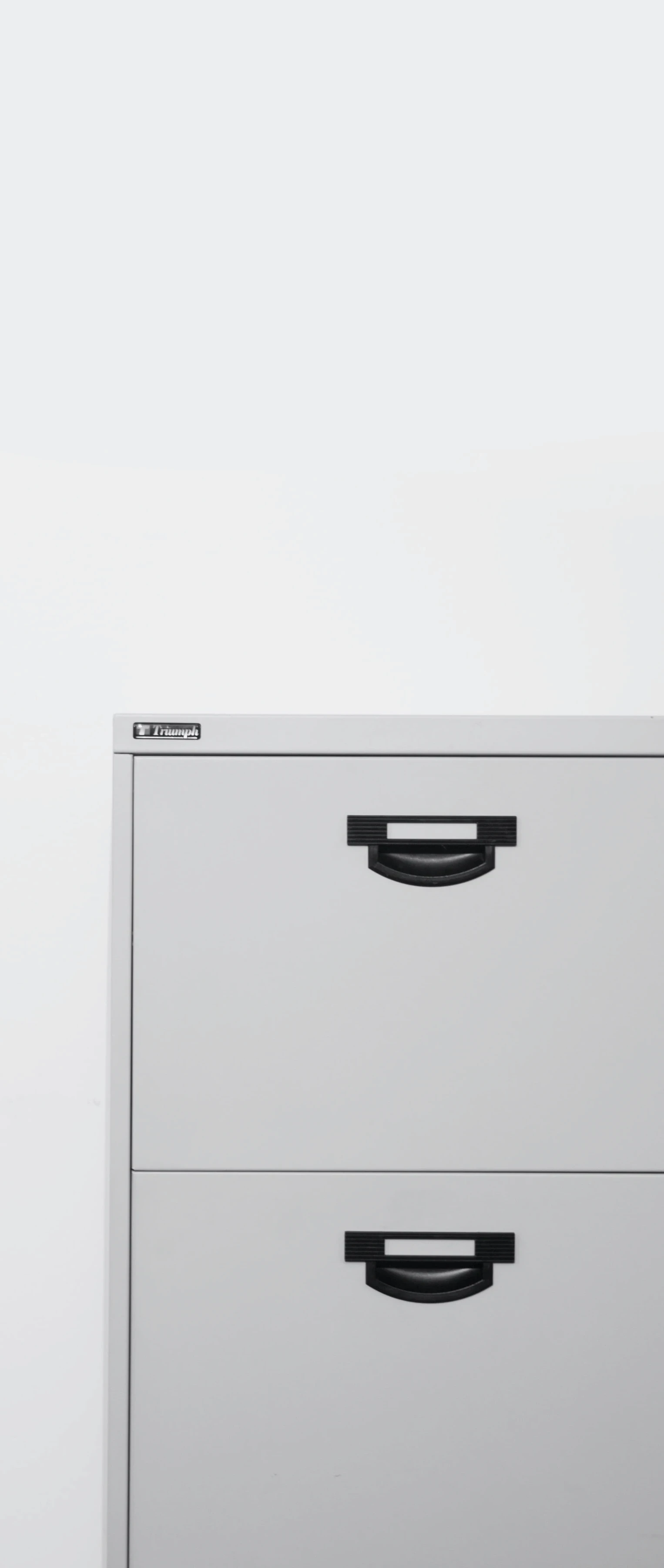 A vertical image featuring the front view of a grey filing cabinet with two drawers, each with a black handle. The cabinet has a label at the top with the text "Triumph," indicating the brand. The background is plain and white, putting the focus on the cabinet.