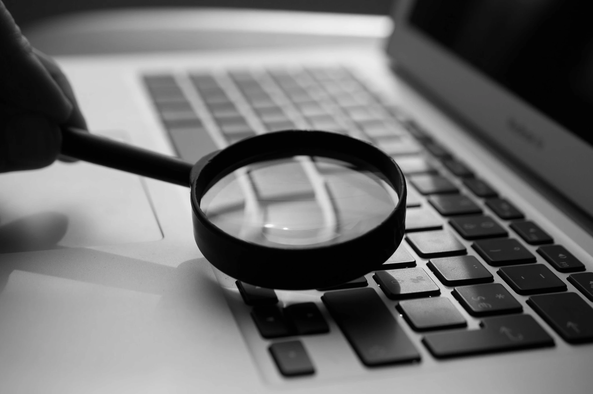 A black and white image depicting a hand holding a magnifying glass over a laptop keyboard, suggesting scrutiny, investigation, or detailed examination. The magnifying glass is focused on the keyboard, blurring the surrounding area and drawing attention to the keys beneath it. The laptop brand is partially visible, indicating a modern and possibly professional context.